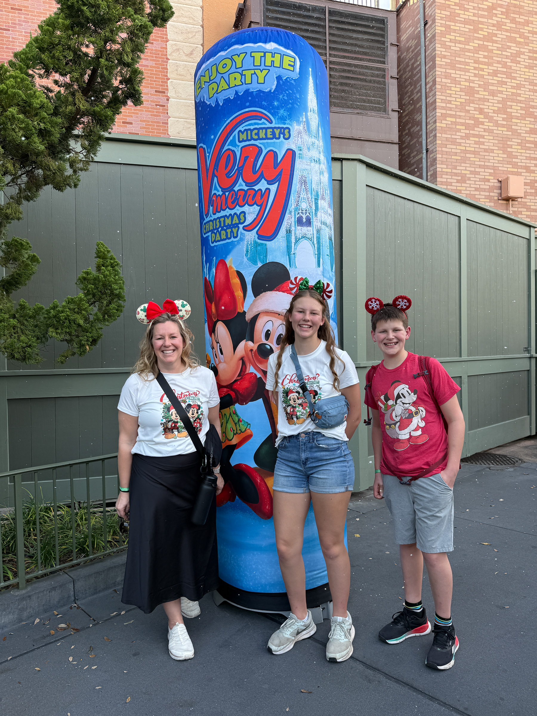 Mom and two teenagers posing beside a Mickey’s Very Merry Christmas Party sign at Magic Kingdom.