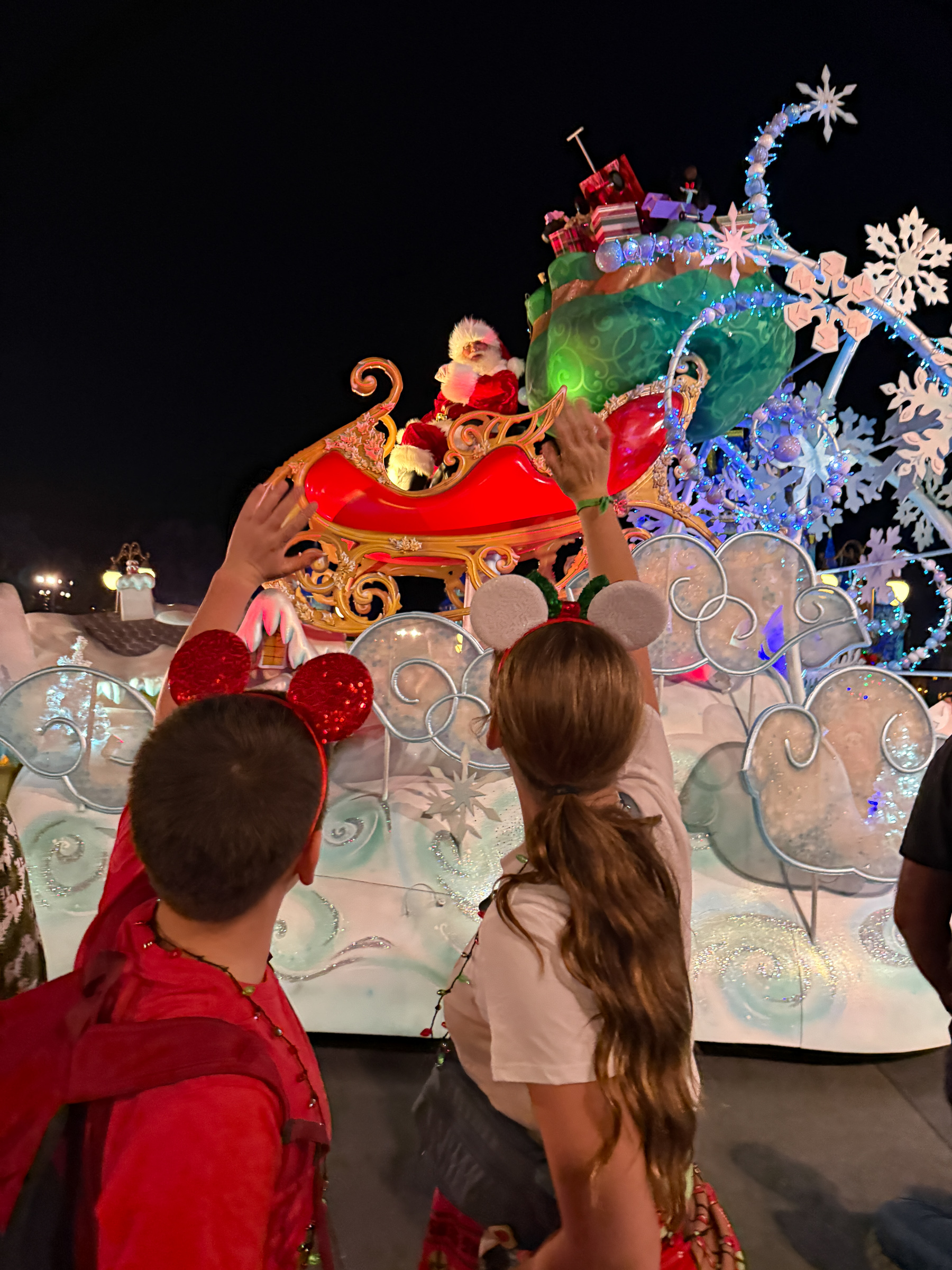 Two teenagers wearing Christmas Mickey ears waving to Santa during Mickey’s Christmas parade at Magic Kingdom.